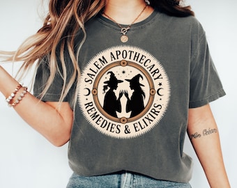 May include: Dark grey t-shirt featuring a circular design with the text "Salem Apothecary Remedies & Elixirs". The design includes silhouettes of two witches in hats. The shirt is a comfortable, casual style.