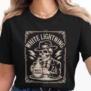 May include: Black t-shirt with a vintage illustration of a skeleton in a suit and fedora, drinking from a jug labelled "White Lightning". The design is framed with decorative elements and lightning bolts.