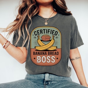 May include: Dark grey t-shirt with a graphic that reads "CERTIFIED BANANA BREAD BOSS." The design features a loaf of bread, two bananas, and a badge-like design in shades of green, orange, and yellow.