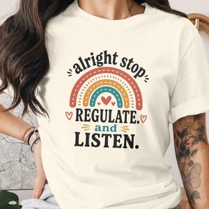 Op de afbeelding: Een crèmekleurig T-shirt met korte mouwen en een grafische print van een veelkleurige regenboog met de tekst "alright stop REGULATE. and LISTEN." in zwarte en blauwgroene letters, geaccentueerd door kleine rode hartjes.