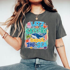 May include: A charcoal gray t-shirt featuring a colorful graphic print with the text "LET'S EXPLORE UNDER THE SEA". The design includes a blue whale, a pink jellyfish, a green sea turtle, a red crab, a seahorse, a starfish, and a clam with a pearl.