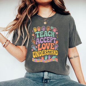 May include: A charcoal gray short-sleeve t-shirt featuring a colorful graphic with the text TEACH ACCEPT LOVE UNDERSTAND. The design includes various hand illustrations, small flowers, hearts, stars, and a checkered pattern at the bottom.