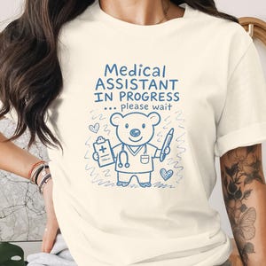 Puede incluir: Una camiseta de manga corta color crema con un gráfico azul de un oso de dibujos animados vestido como profesional médico. El texto "MEDICAL ASSISTANT IN PROGRESS ... please wait" aparece sobre el oso, que sostiene un portapapeles.