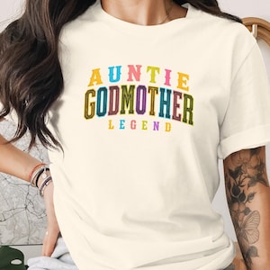 May include: A cream-coloured short-sleeve t-shirt featuring the text AUNTIE GODMOTHER LEGEND printed in a multi-coloured collegiate-style font across the chest. The letters are outlined in various colours including yellow, blue, pink, green, and purple.