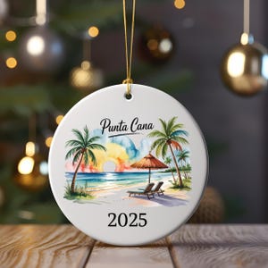 May include: White ceramic Christmas ornament featuring a beach scene with palm trees, a sunset, and the text "Punta Cana 2025." The ornament has a gold string for hanging and is suitable for holiday decor.