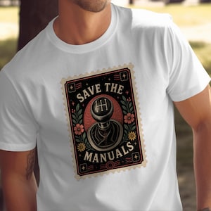 May include: A white short-sleeve t-shirt featuring a rectangular graphic designed like a postage stamp. The graphic displays a black manual transmission gear shifter knob, surrounded by floral motifs and the text "SAVE THE MANUALS".