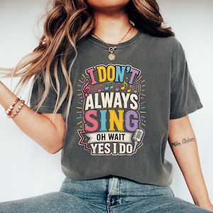 Op de afbeelding: Een antracietgrijs t-shirt met een kleurrijke grafische print met de tekst I DON'T ALWAYS SING OH WAIT YES I DO. Het ontwerp bevat muzieknoten, een microfoonillustratie en levendige letters in de kleuren roze, geel en blauw.
