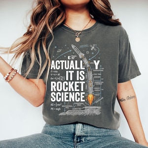 May include: Dark gray t-shirt with the text "ACTUALLY IT IS ROCKET SCIENCE" and a diagram of a rocket with labels. The design includes equations and scientific terms related to rocket science. The shirt is a comfortable, casual wear.
