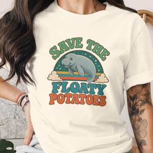 May include: A cream-coloured short-sleeve t-shirt featuring a graphic of a manatee centred within a rainbow-coloured arch. The text "SAVE THE" appears in green above the manatee, and "FLOATY POTATOES" is printed in orange below the graphic.