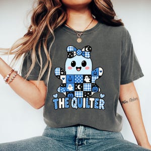 May include: A charcoal grey t-shirt featuring a central graphic of a light blue ghost with a patchwork pattern of blue and black squares, including pumpkins, moons, and stars. The ghost wears a bow and the text THE QUILTER is printed below.