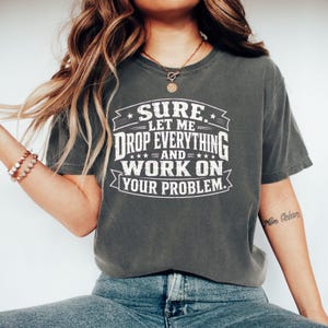 Puede incluir: Una camiseta de manga corta gris carbón con texto blanco desgastado que dice: SURE. LET ME DROP EVERYTHING AND WORK ON YOUR PROBLEM. La camiseta tiene cuello redondo y un ajuste relajado, mostrando un diseño gráfico en la parte delantera.