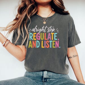 May include: A charcoal grey short-sleeve t-shirt featuring the white cursive text "alright stop." above the colourful block-lettered text "REGULATE. AND LISTEN." in shades of teal, yellow, pink, and orange on the front chest area.