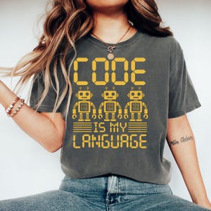 Puede incluir: Camiseta gris oscuro con el texto "CODE IS MY LANGUAGE" en amarillo. El diseño incluye tres figuras de robots con números de código binario. Una camiseta informal para programadores y codificadores.