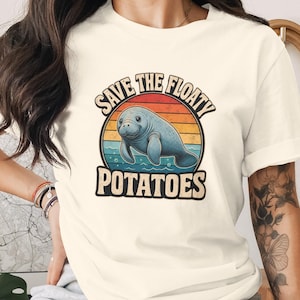 May include: A cream-coloured short-sleeve t-shirt featuring a graphic of a blue manatee swimming in water. The design includes a sunset background with horizontal stripes and the text "SAVE THE FLOATY POTATOES" in a bold, dark font.