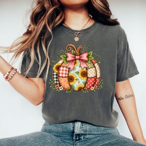 May include: A charcoal grey short-sleeve t-shirt featuring a central graphic of a pumpkin composed of various patchwork patterns including sunflowers, red and white gingham, and polka dots, topped with a large pink bow and green vines.