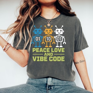Puede incluir: Camiseta gris oscuro con tres robots coloridos y números en código binario. El texto "PEACE LOVE AND VIBE CODE" está impreso debajo de los robots en verde y amarillo.