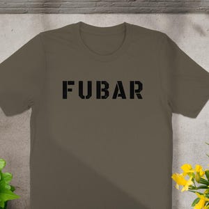 Puede incluir: Una camiseta de manga corta y cuello redondo en color verde oliva sólido. En el centro del pecho aparece la palabra FUBAR impresa en una fuente negra en negrita de estilo plantilla, con una textura desgastada sobre las letras.