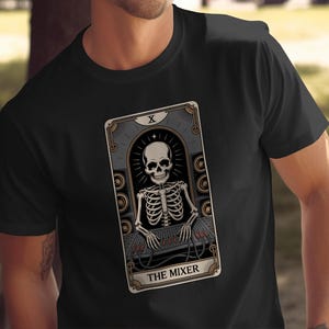 May include: Black t-shirt with a tarot card design. The card depicts a skeleton at a sound mixer, labelled "The Mixer". The design has a vintage look with grey, beige, and gold tones. The image is centrally placed on the shirt.