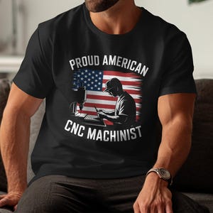 May include: Black t-shirt with the text "Proud American CNC Machinist" above an illustration of a machinist working with a CNC machine, set against a backdrop of the American flag.