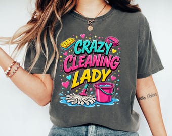 May include: A charcoal gray t-shirt featuring a colorful graphic with the text "CRAZY CLEANING LADY" in bold pink and yellow letters. The design includes a yellow sponge, a pink bucket, and a white mop surrounded by small hearts and dots.