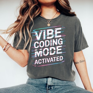 May include: Dark grey t-shirt with the text "VIBE CODING MODE ACTIVATED" in a glitch effect design. The text is white with teal, pink, and blue horizontal lines. The shirt is a casual style.