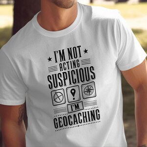 May include: A white short-sleeved t-shirt featuring black text that reads I'M NOT ACTING SUSPICIOUS I'M GEOCACHING. The design includes three square icons depicting a compass, a location pin, and a star, accented by horizontal lines and stars.