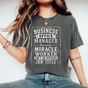 Peut inclure: Un t-shirt gris anthracite à manches courtes avec un texte blanc vieilli indiquant : BUSINESS OFFICE MANAGER because MIRACLE WORKER ISN'T AN OFFICIAL JOB TITLE. Le motif est encadré par une bordure rectangulaire avec des lignes et des étoiles.
