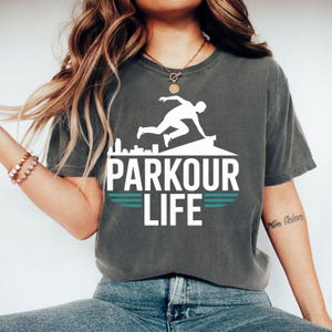 May include: Dark gray t-shirt with the words "PARKOUR LIFE" in white block letters. A white silhouette of a person doing parkour is above the text. Two horizontal teal lines are below the text. The shirt is a comfortable, casual style.