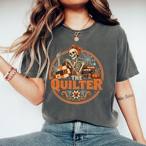May include: A charcoal grey t-shirt featuring a graphic of a skeleton wearing a polka dot headscarf and sewing a quilt. The design includes spiderwebs, a stack of fabric, a pumpkin, and the text THE QUILTER in bold orange letters within a circle.