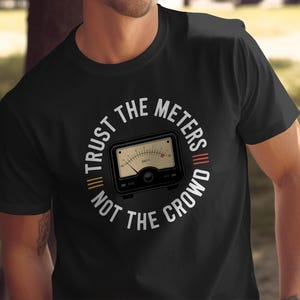 May include: Black t-shirt featuring a circular graphic with the text "TRUST THE METERS NOT THE CROWD." The graphic includes a vintage-style meter with a needle and gauge. The shirt is a crew neck style.