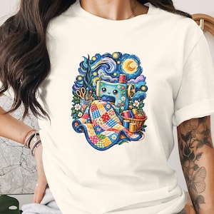 May include: A cream-coloured short-sleeve t-shirt featuring a colourful graphic print of a blue sewing machine stitching a multi-coloured patchwork quilt. The design includes a starry night sky background, scissors, spools of thread, and flowers.