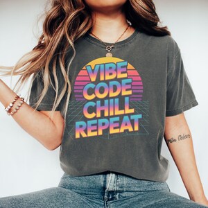 Puede incluir: Camiseta gris oscuro con un diseño retro. El diseño presenta las palabras "VIBE CODE CHILL REPEAT" en un degradado de colores, sobre un fondo estilo puesta de sol. La camiseta es de estilo casual y cómodo.