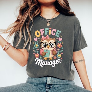Op de afbeelding: Een antracietgrijs t-shirt met een afbeelding van een cartoonuil met een bril en een roze strik, die een klembord en een pen vasthoudt. De tekst "OFFICE Manager" staat in witte en kleurrijke letters, omringd door kleine hartjes en bloemen.