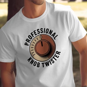 Op de afbeelding: Wit T-shirt met een cirkelvormige afbeelding. De afbeelding toont "PROFESSIONAL KNOB TWISTER" rond een bruine wijzerplaat met een naald. Het shirt met korte mouwen wordt door een persoon gedragen.