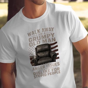 Może przedstawiać: Biały t-shirt z krótkim rękawem z grafiką czarnego skórzanego fotela na tle wyblakłej amerykańskiej flagi. Tekst głosi: WALK AWAY I AM A GRUMPY OLD MAN I HAVE ANGER ISSUES AND A SERIOUS DISLIKE FOR STUPID PEOPLE.