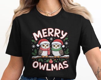 May include: Black t-shirt featuring a festive design with two owls wearing Santa hats and scarves, perched on a branch. The text "MERRY OWLMAS" is displayed above and below the owls, with Christmas-themed accents.
