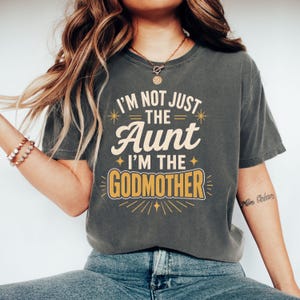 Op de afbeelding: Een antracietgrijs t-shirt met korte mouwen en de crème en gele tekst I'M NOT JUST THE AUNT I'M THE GODMOTHER. Het ontwerp bevat decoratieve ster- en lijnaccenten rondom de dikgedrukte letters op de voorzijde van het kledingstuk.