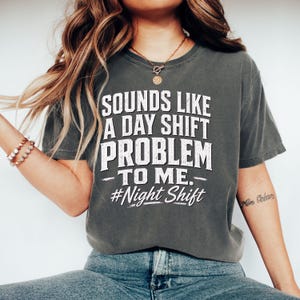 Puede incluir: Una camiseta de manga corta gris carbón con texto blanco desgastado que dice "SOUNDS LIKE A DAY SHIFT PROBLEM TO ME." Debajo del texto principal, el hashtag "#Night Shift" está impreso en una fuente cursiva blanca en el pecho.