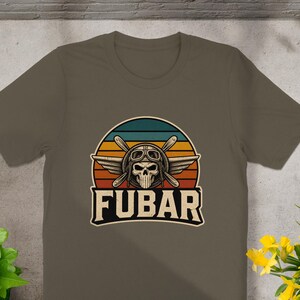Puede incluir: Una camiseta de manga corta de color verde oliva oscuro con un estampado gráfico de una calavera con gafas de aviador y casco, flanqueada por dos hélices de avión. Detrás de la calavera hay un círculo a rayas con colores de atardecer.