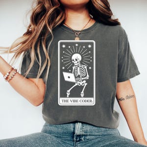 May include: Dark grey t-shirt with a white graphic of a skeleton using a laptop, framed by a rectangle with starburst and star accents. The text "THE VIBE CODER." is printed below the skeleton. The shirt is a casual wear item.