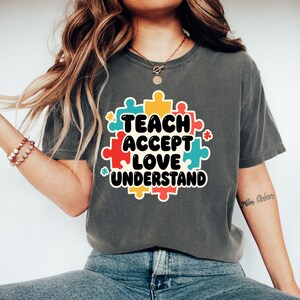 May include: A charcoal grey short-sleeve t-shirt featuring a central graphic of colourful puzzle pieces in red, yellow, and blue. The text "TEACH ACCEPT LOVE UNDERSTAND" is printed in bold black letters across the centre of the puzzle design.