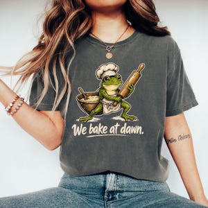 May include: A charcoal gray t-shirt featuring a graphic of a green frog wearing a white chef hat and apron, holding a wooden rolling pin next to a mixing bowl. The text "We bake at dawn." is printed in white below the illustration of the frog.