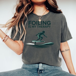 Peut inclure: T-shirt gris foncé avec l'inscription "FOILING IS MY THERAPY" en vert. Le t-shirt présente un graphisme vert d'une personne faisant du foil sur l'eau. Le t-shirt est de style décontracté et confortable.