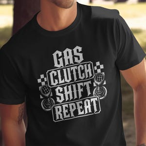 Op de afbeelding: Een zwart t-shirt met korte mouwen en een witte grafische print met de tekst GAS CLUTCH SHIFT REPEAT. Het ontwerp bevat twee handmatige versnellingspookknoppen en een geblokt vlagpatroon aan de zijkanten van de dikke, gestileerde letters.