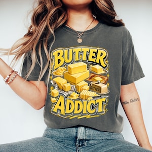 May include: Dark grey t-shirt with a yellow graphic that reads "BUTTER ADDICT." The graphic features illustrations of butter in various forms, including blocks, pats, and a butter dish. The shirt is a casual style.