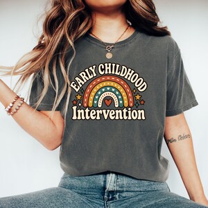 May include: A charcoal gray short-sleeve t-shirt featuring a graphic print of a multi-colored rainbow with small hearts and stars. The text "EARLY CHILDHOOD Intervention" is printed in a curved, bold, cream-colored font above and below the rainbow.