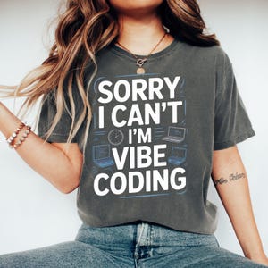 May include: Dark gray t-shirt with the text "SORRY I CAN'T I'M VIBE CODING" in white and blue. The design includes laptop and clock graphics. The shirt is a casual wear item.