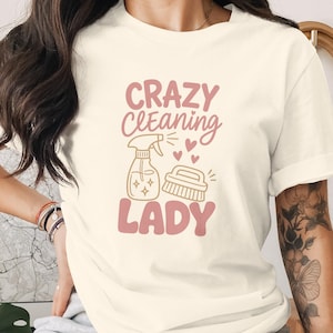 Könnte beinhalten: Ein cremefarbenes T-Shirt mit dem Text CRAZY CLEANING LADY in einer kräftigen, altrosa Schrift. Unter dem Text befindet sich eine Strichzeichnung einer Sprühflasche und einer Scheuerbürste in hellem Beige, akzentuiert durch drei kleine rosa Herzen.