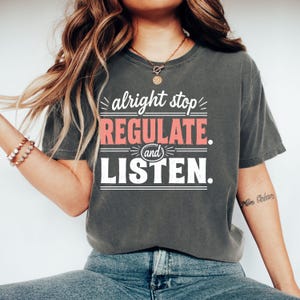 Puede incluir: Una camiseta de manga corta en color gris carbón con texto en blanco y salmón que dice alright stop REGULATE and LISTEN. La prenda tiene cuello redondo y un corte relajado, con el gráfico centrado en la zona del pecho de la camiseta.
