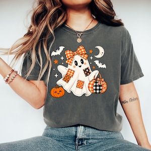 May include: A charcoal grey t-shirt featuring a graphic of a white ghost with orange and black plaid patches, a matching bow, and a pumpkin. The design includes small bats, stars, and a crescent moon, creating a festive seasonal aesthetic.